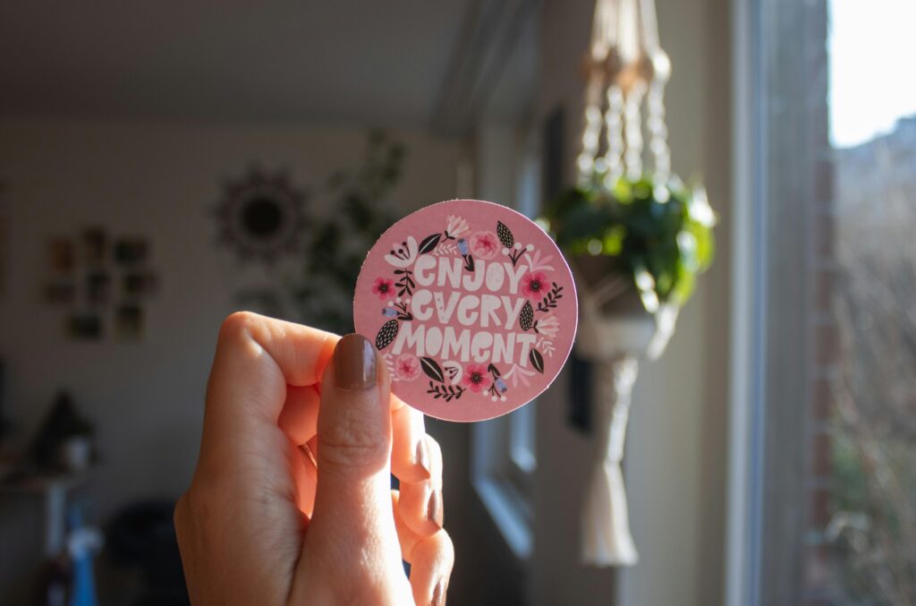 A hand holding a decorative sticker with 'Enjoy Every Moment' text in a cozy indoor setting.